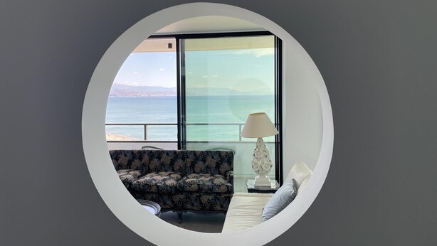 A round window with a view of the sea and distant mountains. Inside, there is a sofa with a floral pattern, a white lamp, and a small table with a decorative item.