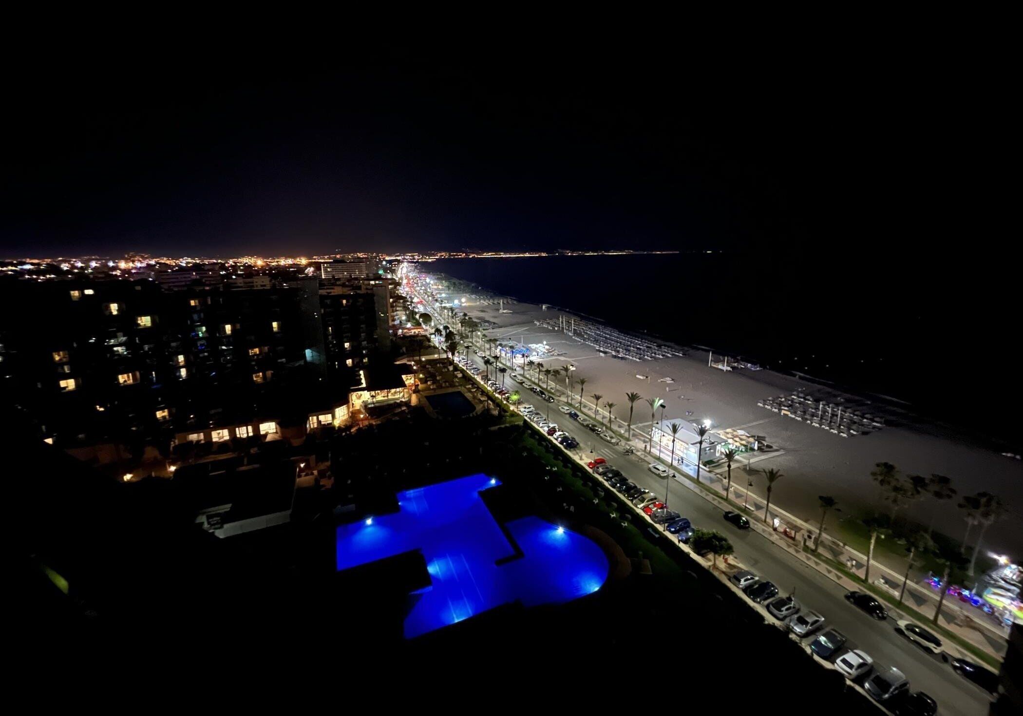 A nighttime aerial view of a coastal hotel with a swimming pool, street lighting, and a view of the ocean.