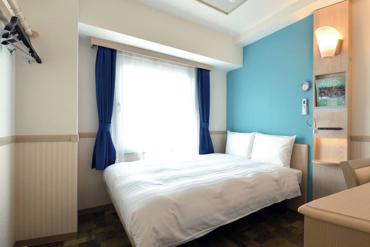 Habitació d'hotel amb un llit, un escriptori, una finestra amb cortines, un rellotge i una làmpada de paret.
