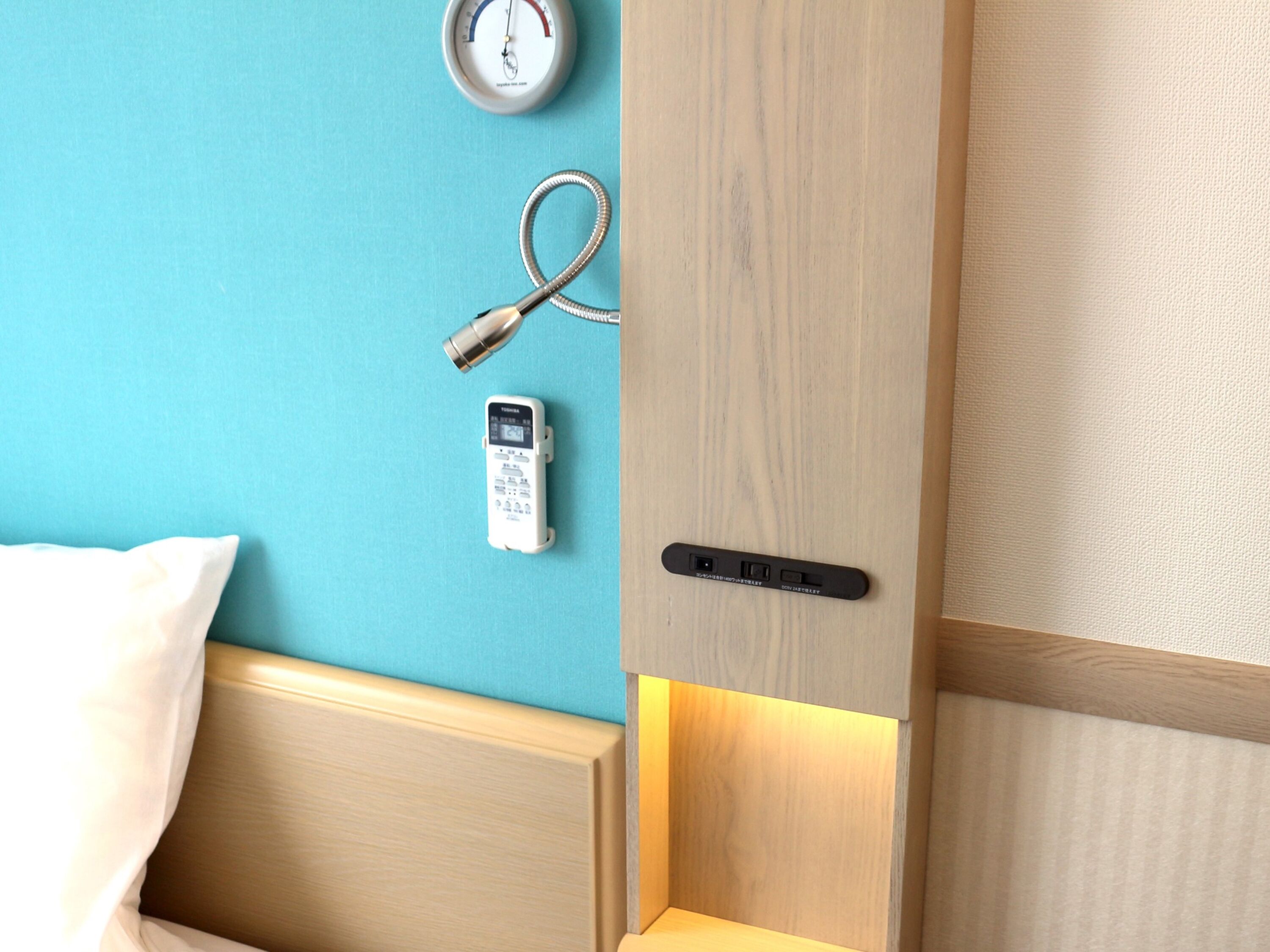 A hotel room with a wooden headboard, a bed with white bedding, a wall-mounted clock, a telephone, and a door with a handle.