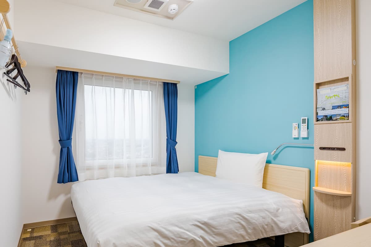 Habitació d'hotel amb un llit, una taula de nit, una finestra amb cortines, un panell de control fixat a la paret i una estant fixa a la paret amb un llibre.