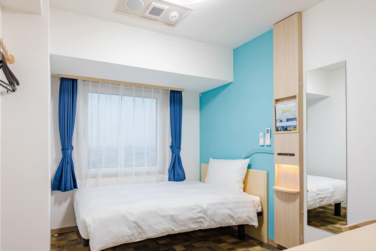 Habitació d'hotel amb un llit, una finestra amb cortines blaves, un mirall i un panell de control fixat a la paret.