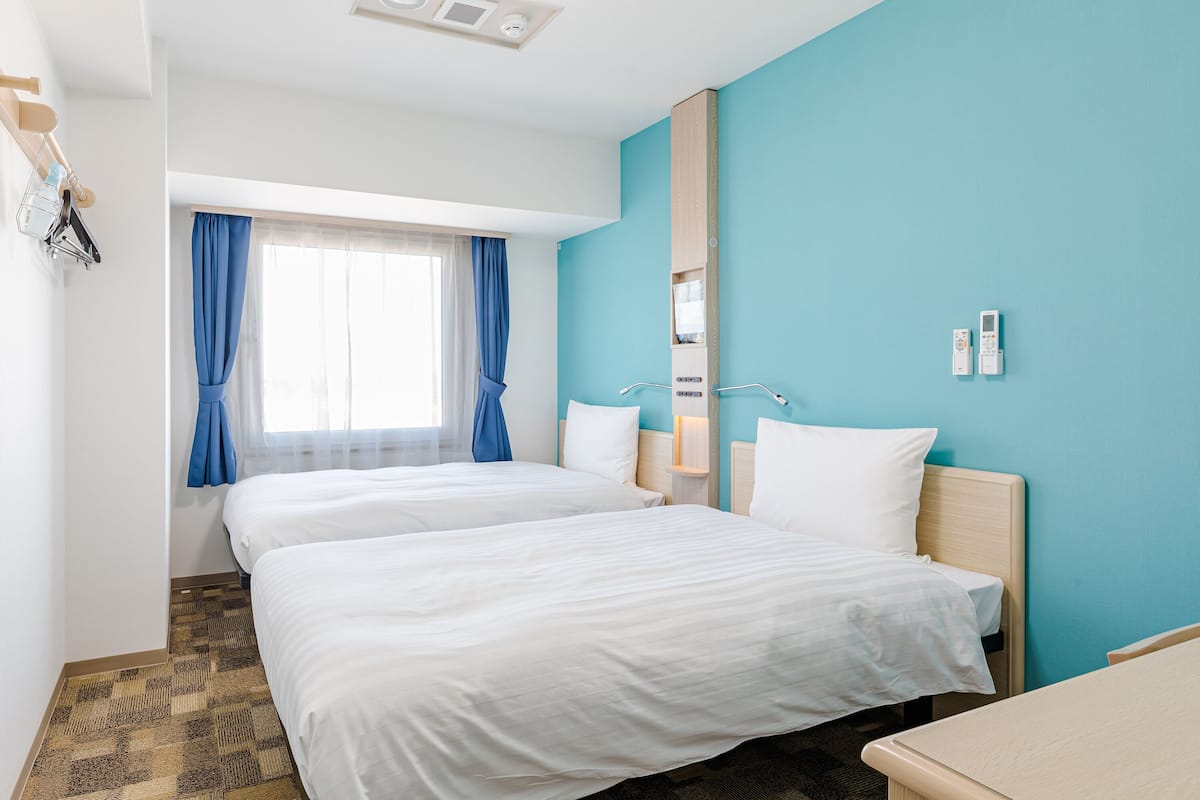 Habitació d'hotel amb dos llits, una finestra amb cortines blaves, una taula de nit i un panell de control fixat a la paret.