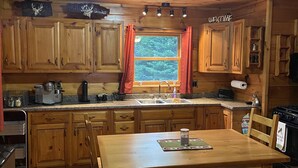 Cocina de cabaña de madera con mesa y banco, una ventana con cortinas y un cartel con la palabra 'WELCOME' sobre la puerta.