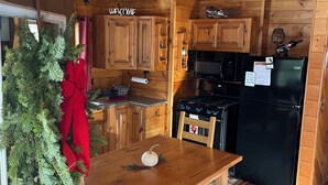 Interior de una cabaña de madera con una cocina, una mesa de comedor con bancos y un árbol de Navidad con un lazo rojo.