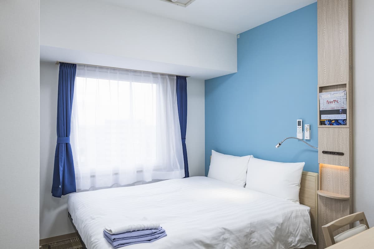 A hotel room with a bed, a desk, a chair, a window with curtains, a wall-mounted control panel, and a small shelf with items.