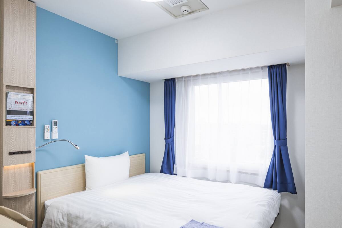 A hotel room with a large bed, a bedside table, a wall-mounted control panel, and a window with blue curtains.