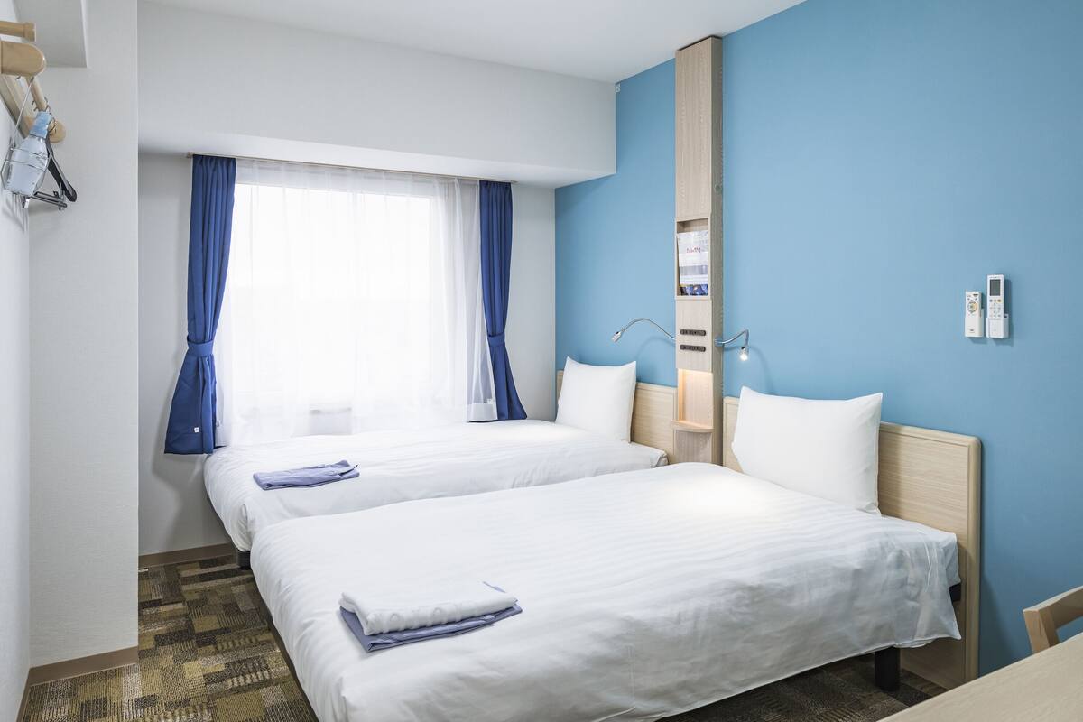 A hotel room with two beds, a large window with curtains, a bedside table, a lamp, and a wall-mounted thermostat.