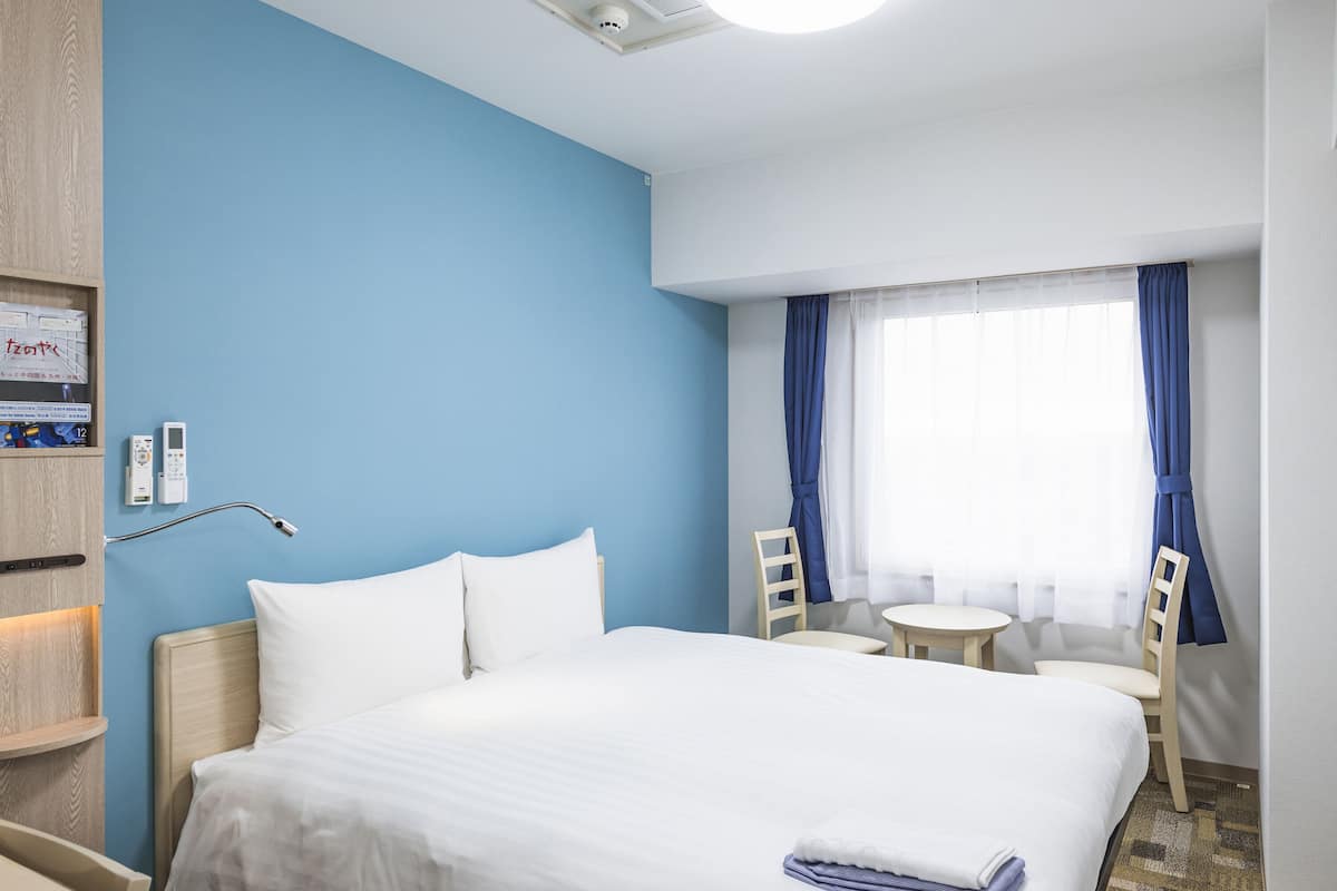 A hotel room with a large bed, a bedside table, a chair, a small table, and a window with curtains.