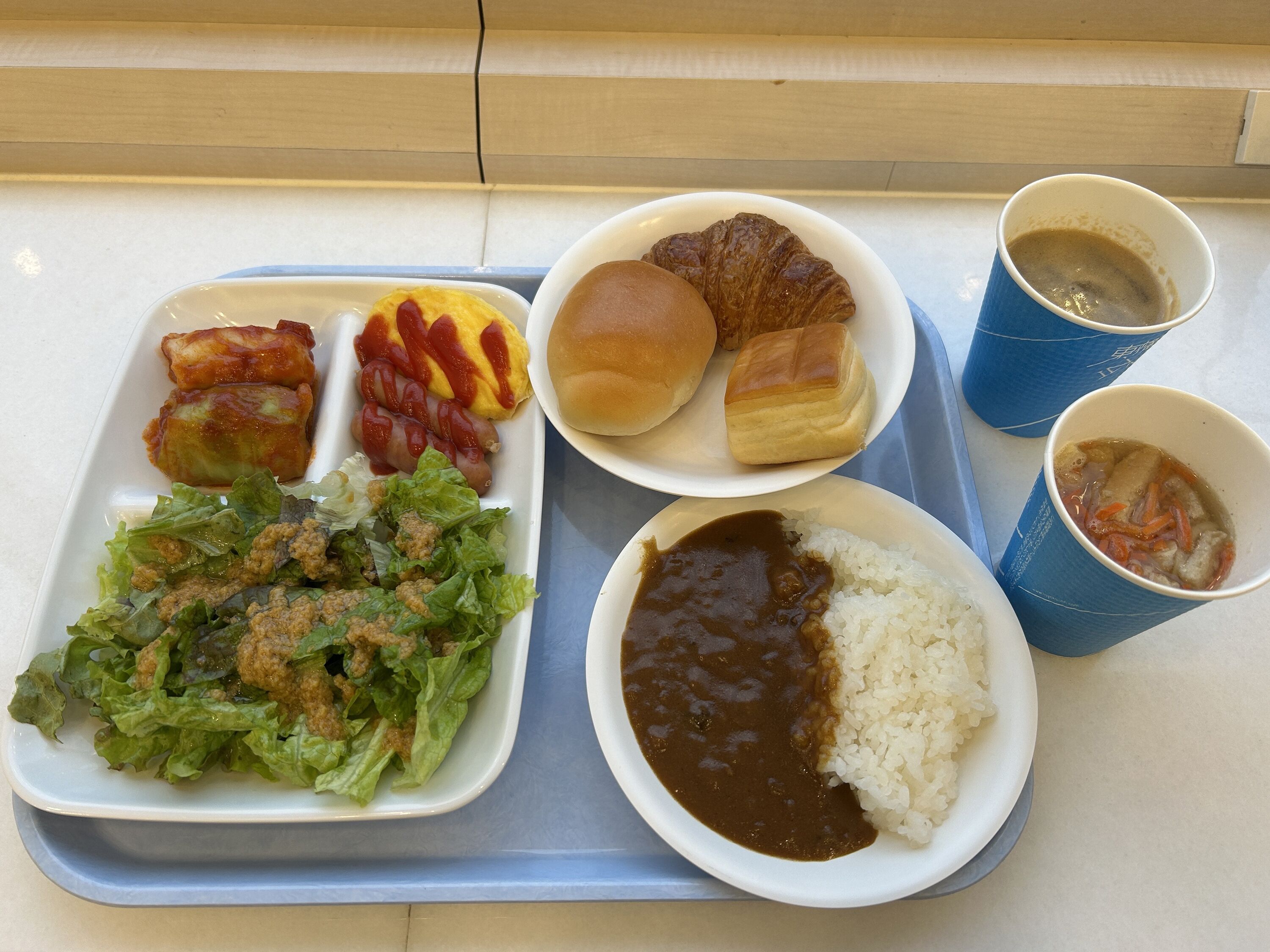 A tray with food items including a salad, rice with beans, sausages, a croissant, and two cups of coffee.