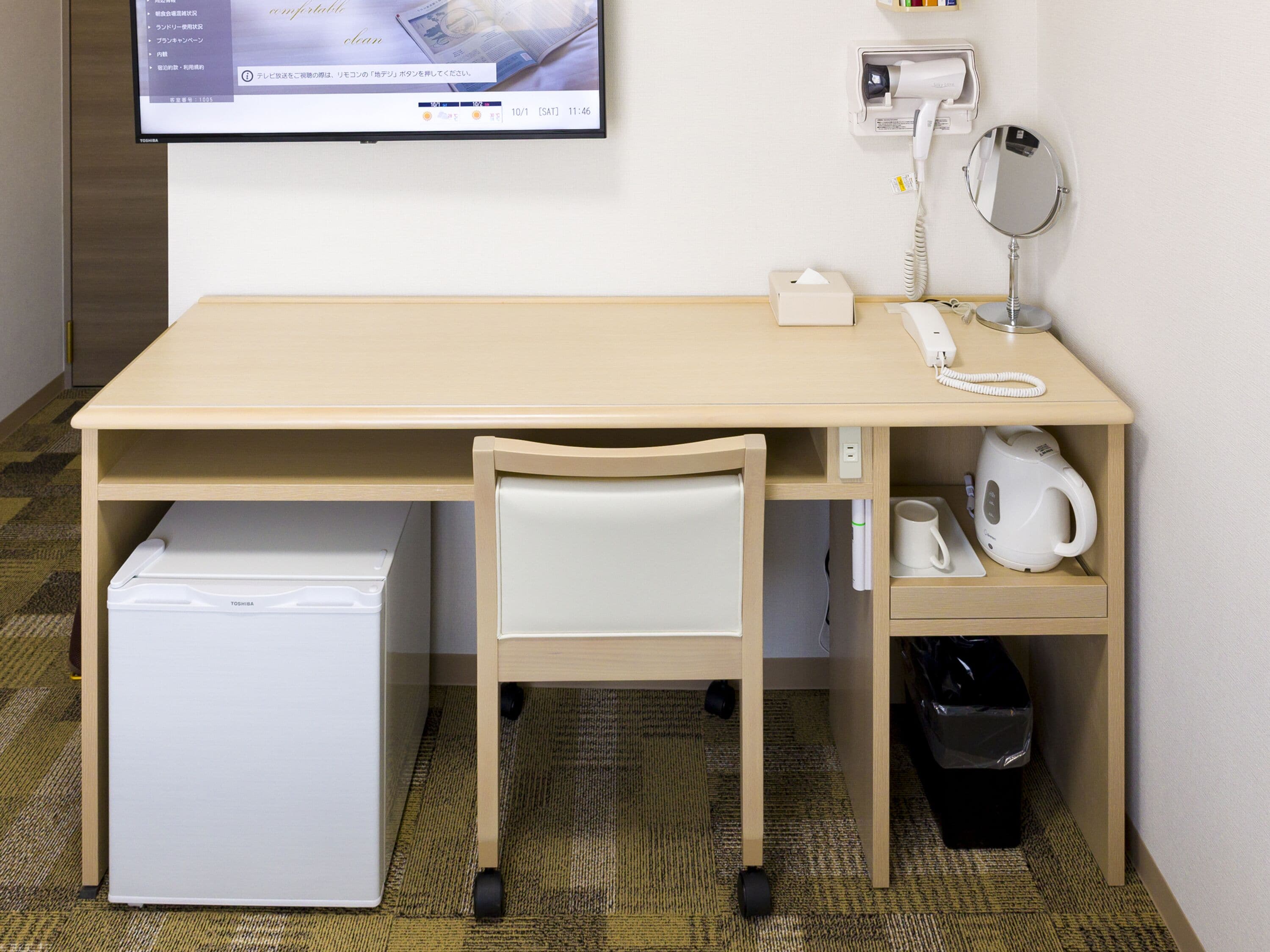 Habitación de hotel compacta con escritorio de madera, una silla, refrigerador, teléfono, espejo, hervidor de agua y un cesto de basura.