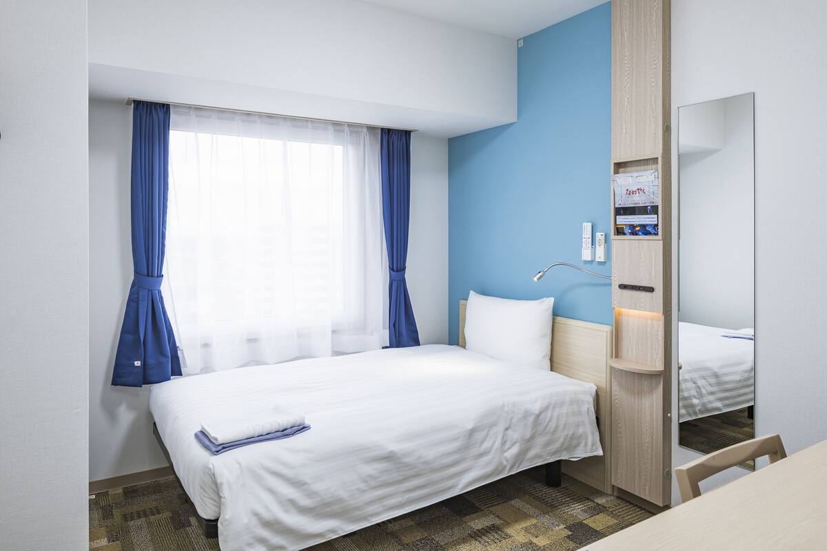 A hotel room with a bed, a desk, a chair, a mirror, a window with curtains, and a wall-mounted shelf with items.