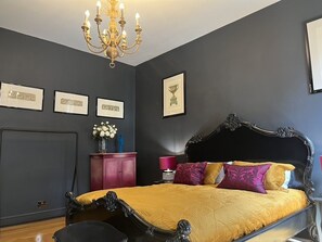 Ein Schlafzimmer mit dunkel gestrichener Wand, einem Bett mit aufwendigem Kopfteil, einer Chandelüster, gerahmten Bildern und einem pinken Schrank.