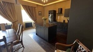 A modern kitchen with dark cabinetry, a central island, and a dining area with a wooden table and chairs. - Luxury flat 2 mins from tram stop in the 4th coolest neighbourhood in the world! (Edinburgh)
