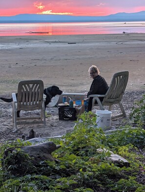 Eine Person sitzt an einem Tisch mit einem Hund, draußen mit Blick auf ein Gewässer und ferne Berge.