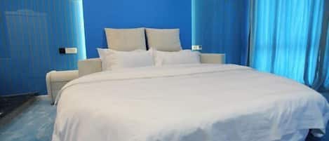A large bed with white bedding, a nightstand, and a wall with blue curtains.