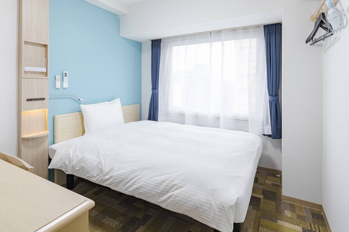 Une chambre d’hôtel comprenant un lit, un bureau, une armoire, une fenêtre avec des rideaux et une lampe fixée au mur.