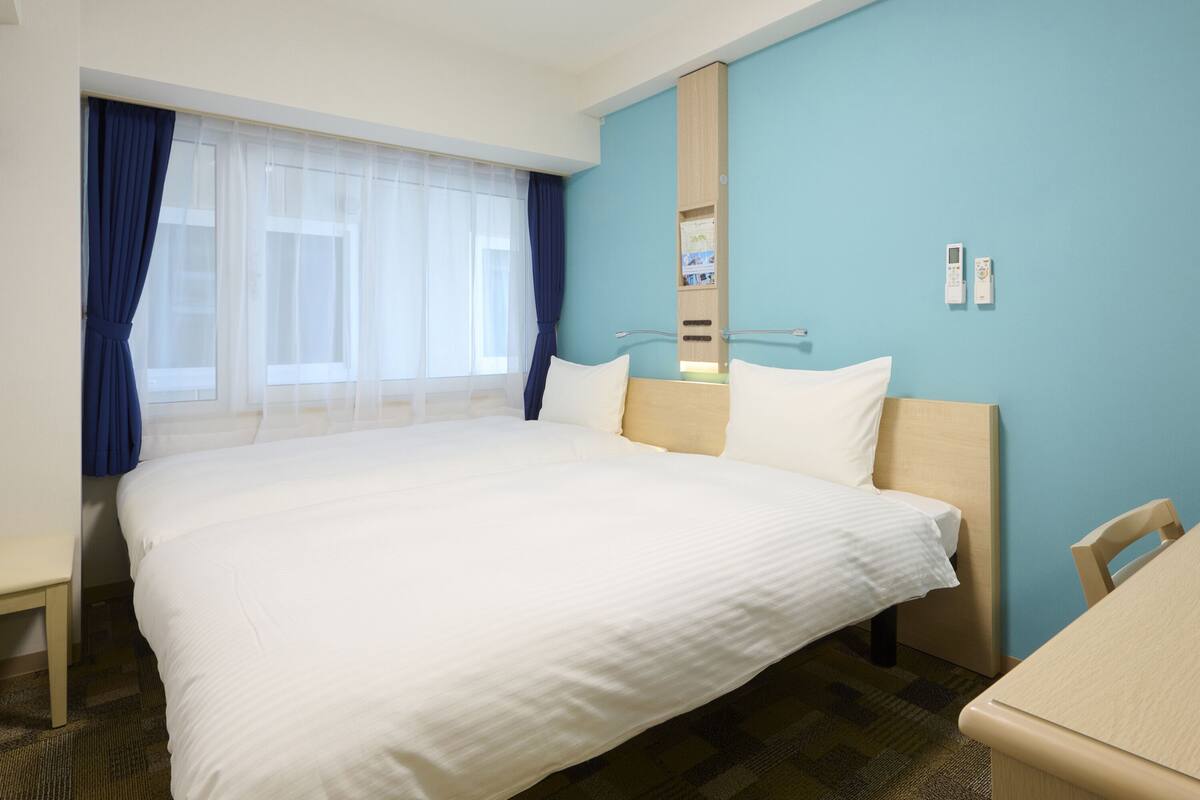 Une chambre d’hôtel avec un grand lit, un bureau et une chaise. Il y a une fenêtre avec des rideaux, un miroir et un thermostat fixé au mur.