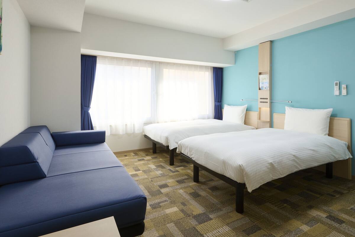 Une chambre d’hôtel avec deux lits, un canapé, une fenêtre avec des rideaux, un luminaire au plafond et un téléphone fixé au mur.