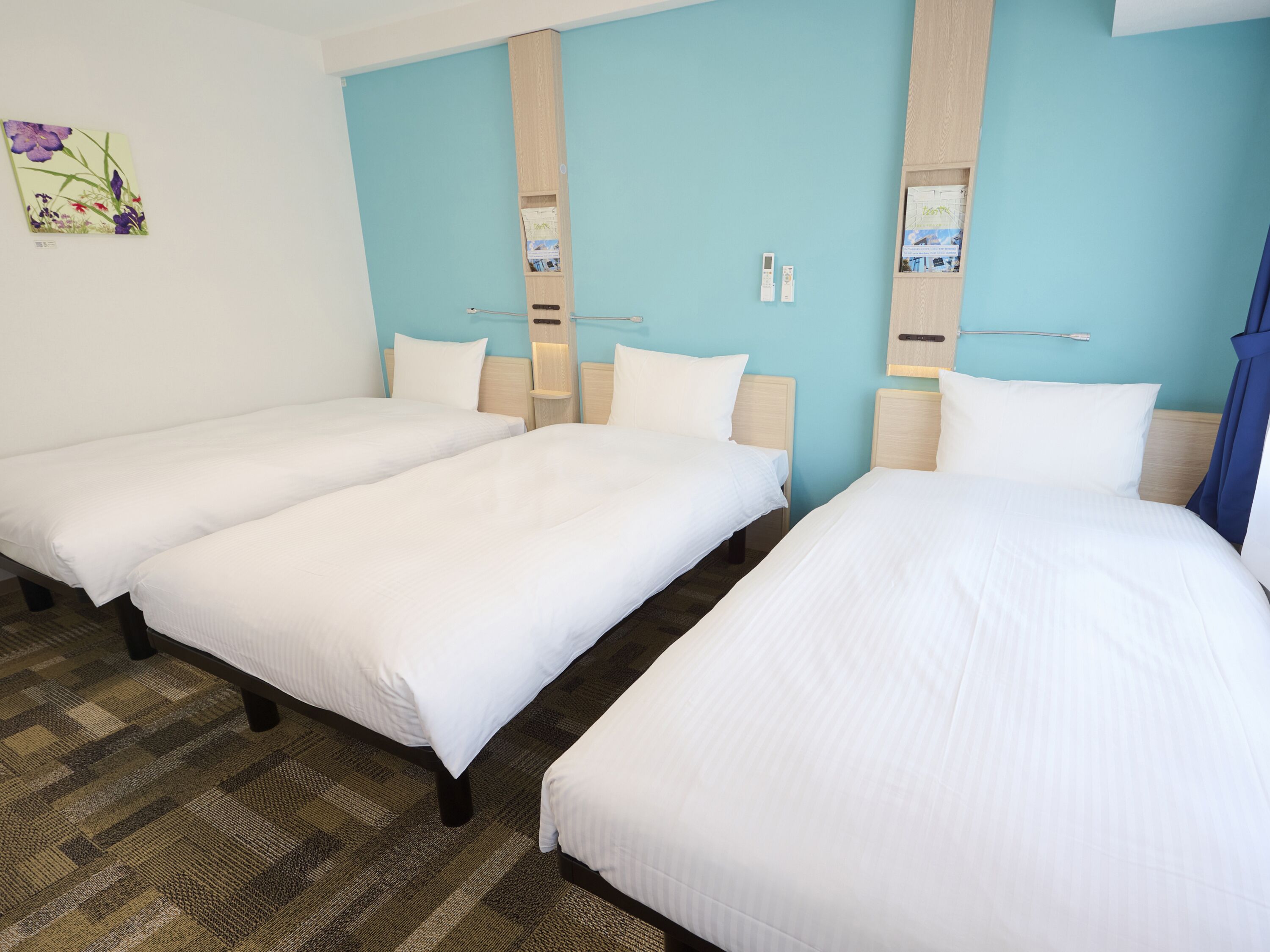 A hotel room with three single beds, a wooden headboard, a blue accent wall, and a small painting of flowers.