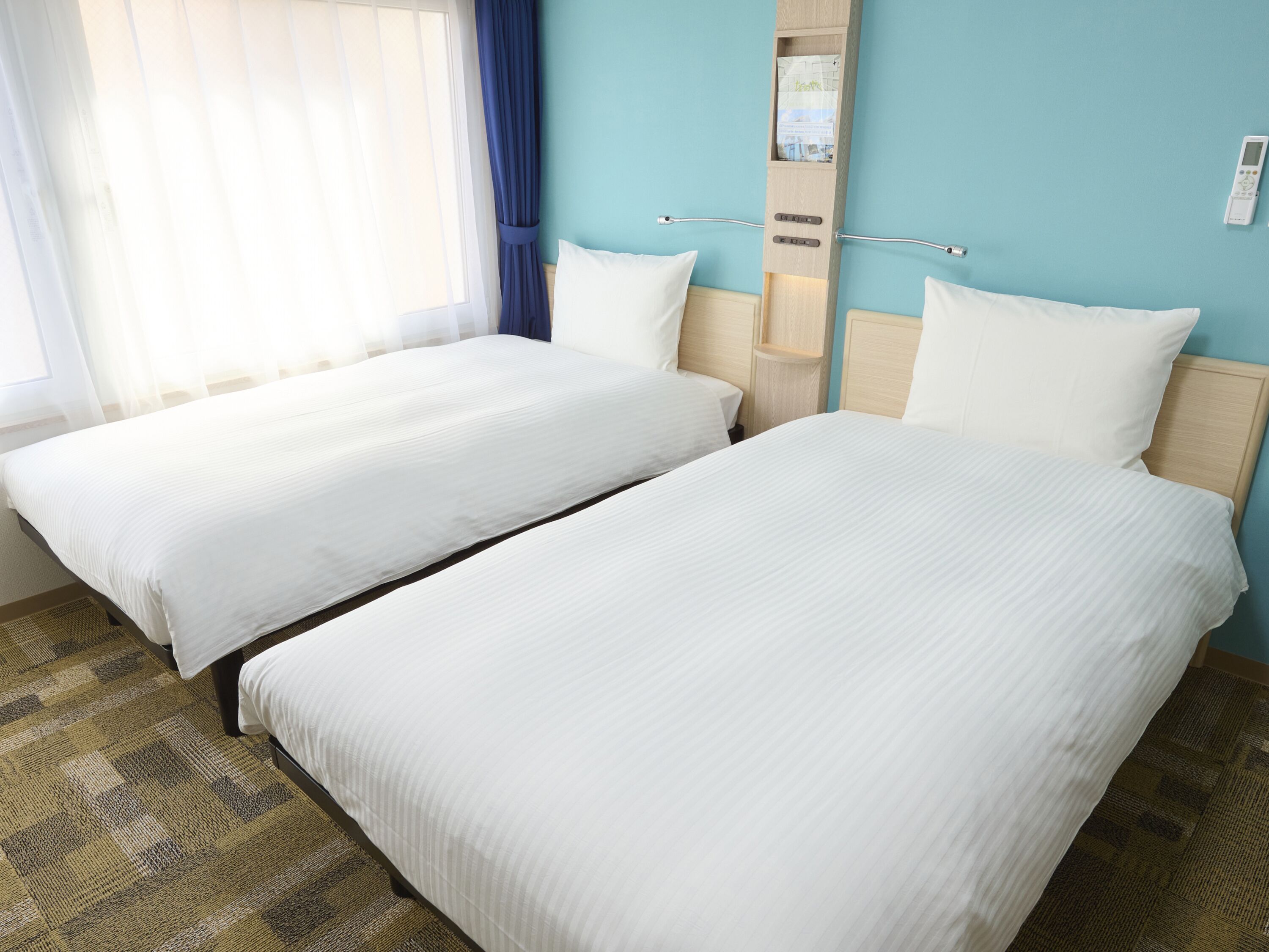A hotel room with two single beds, a wooden headboard, a small shelf, and a window with curtains.