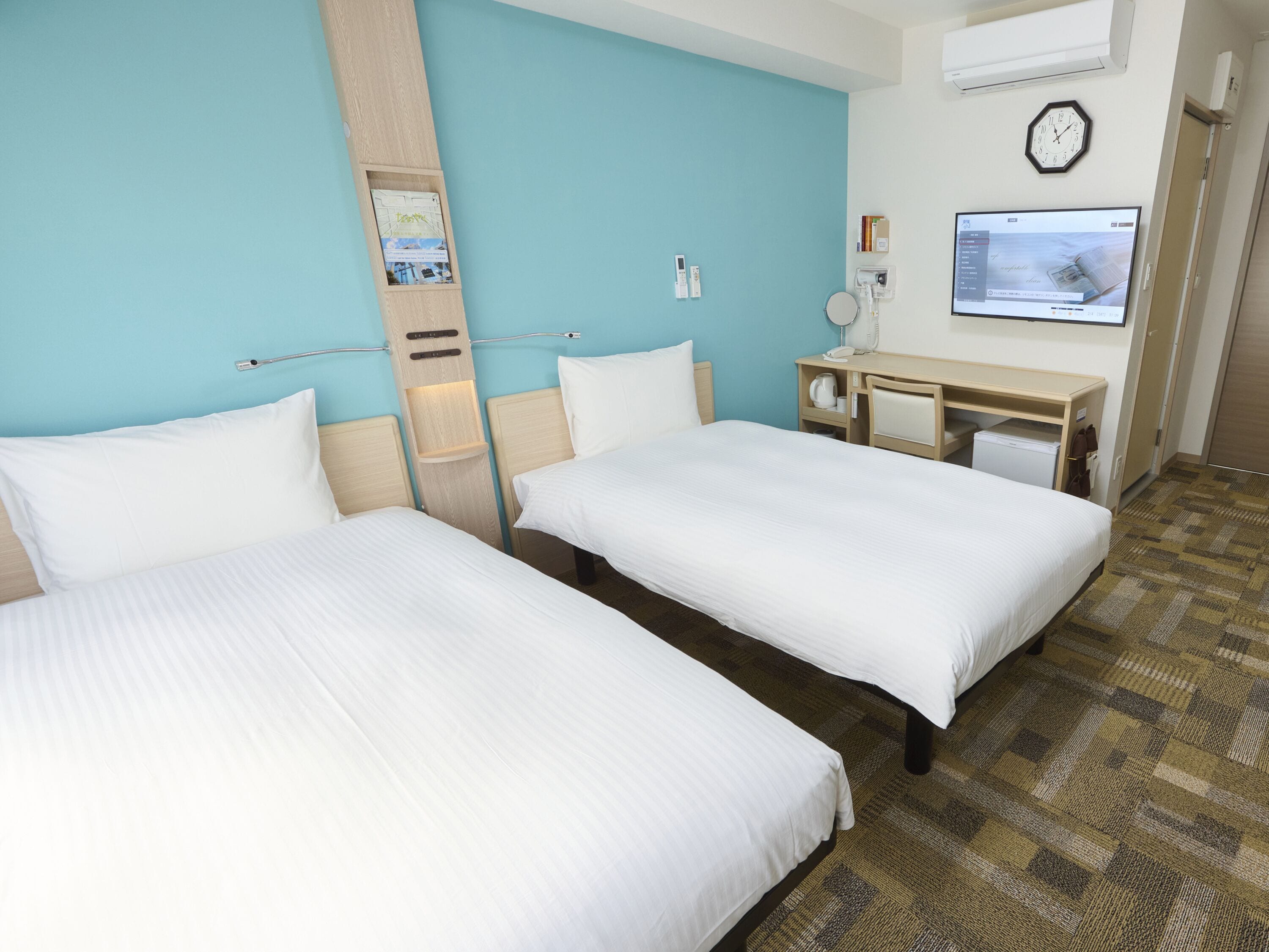 A hotel room with two single beds, a desk with a chair, a television, and a clock on the wall.