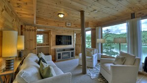 A wooden cabin interior with a TV, white sofas, and a large window overlooking a lake. - The Blue Canoe | Cozy Lakefront Getaway in Blue Ridge | Rustic and Elegant  (Blue Ridge)