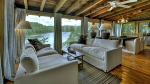 A spacious living area with a large window overlooking a lake, a ceiling fan, and a dining area with a table and chairs. - The Blue Canoe | Cozy Lakefront Getaway in Blue Ridge | Rustic and Elegant  (Blue Ridge)
