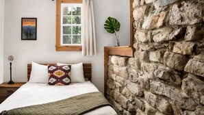 Ein Schlafzimmer mit Steinwand, einem Bett mit gemustertem Kopfkissen, einem Fenster mit Vorhängen und einem Poster an der Wand.