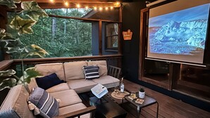 Ein gemütlicher Außenbereich mit einer Couch, einem kleinen Tisch und einem Fernseher, der eine Berglandschaft zeigt.