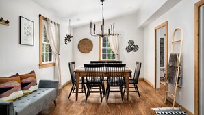 Ein Essbereich mit Holztisch, schwarzen Stühlen und einer Pendelleuchte. Es gibt ein Sofa mit bunten Kissen und Holzdielen.