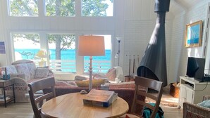 A cozy living room with a round wooden table, comfortable seating, a large window with a view of the ocean, and a chimney. - Sunset Serenity - INCREDIBLE HOME ON BLUFF OVERLOOKING LAKE MICHIGAN! (Frankfort)