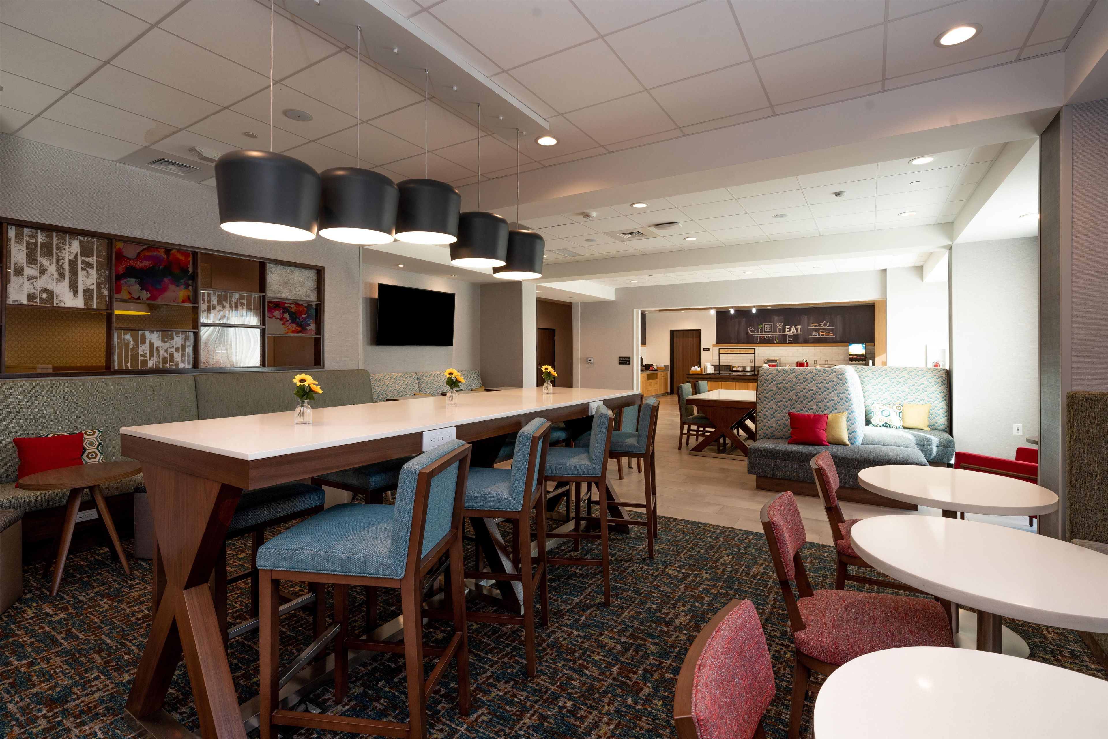 A hotel lobby with a long communal table, several chairs, a sofa, and a television mounted on the wall.
