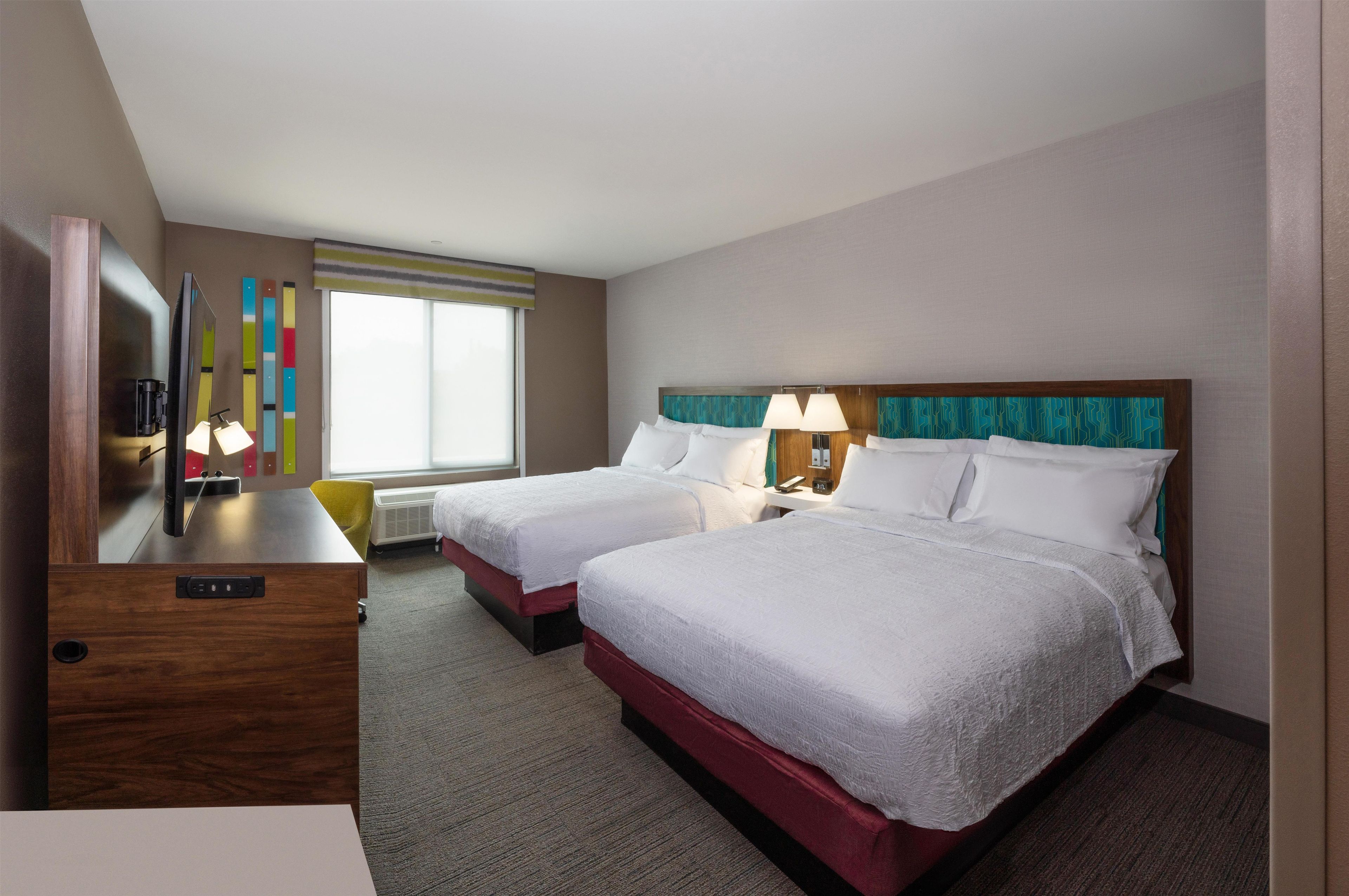 A hotel room with two beds, a desk, a chair, a TV, and a window with blinds.