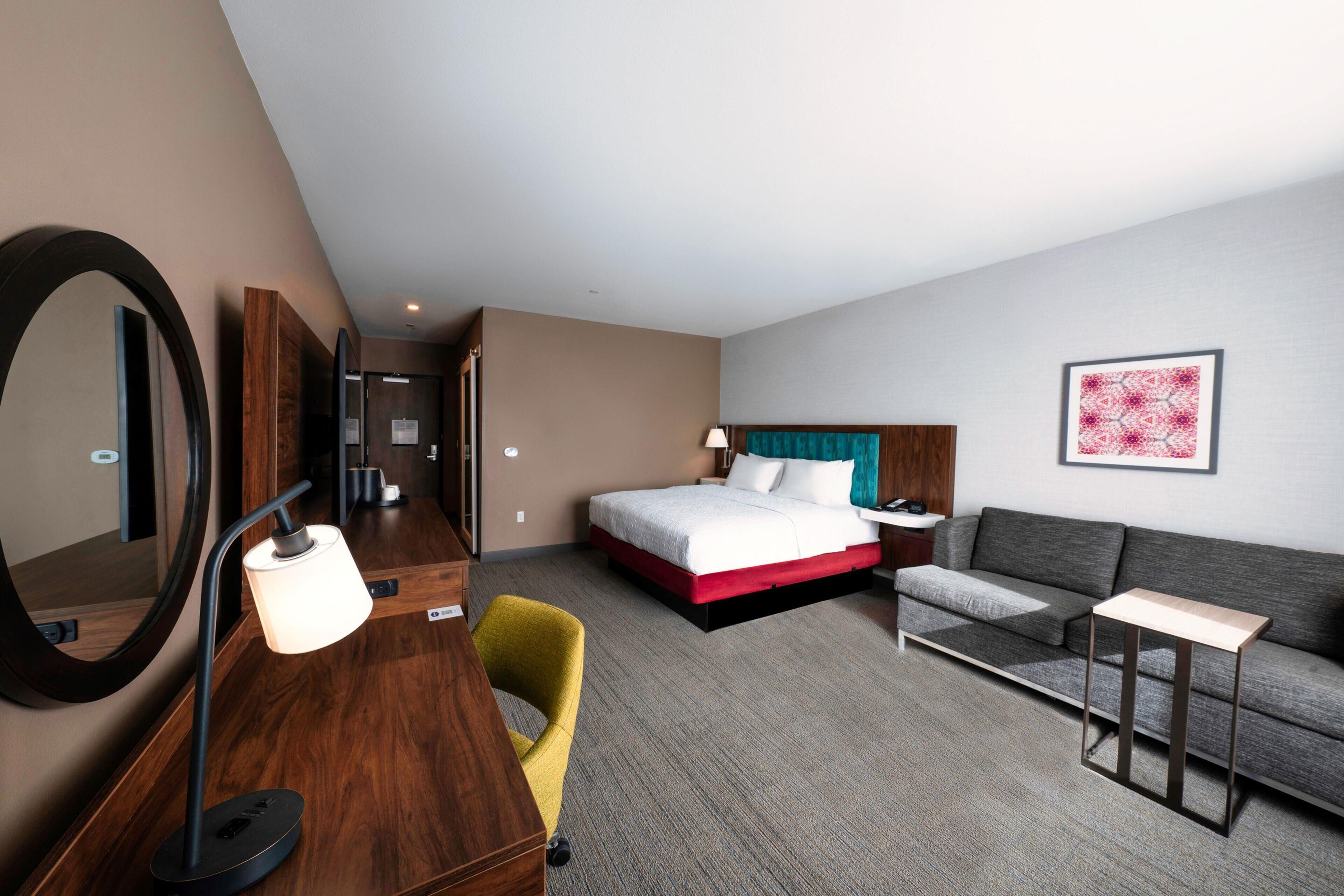 A hotel room with a bed, a sofa, a desk, a chair, a lamp, a mirror, and a framed picture on the wall.