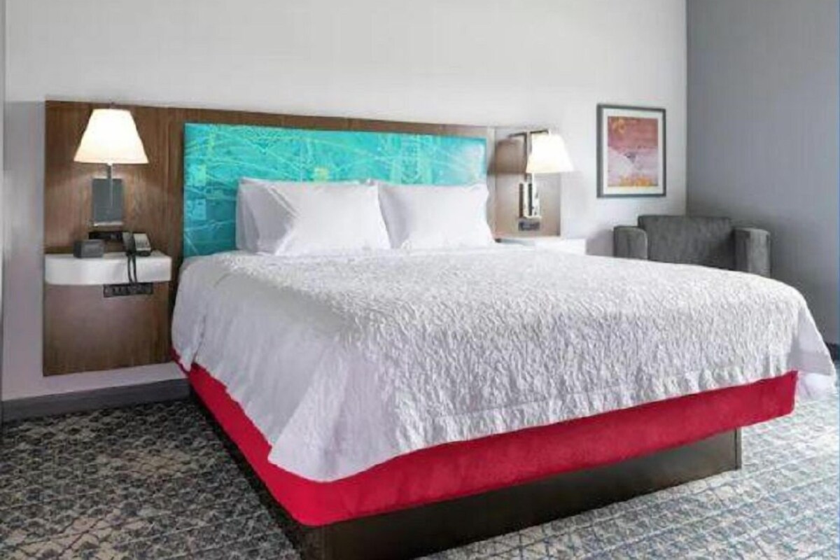 Una habitación de hotel con una cama, dos lámparas de noche, una mesita de noche, una silla y un cuadro colgado en la pared.