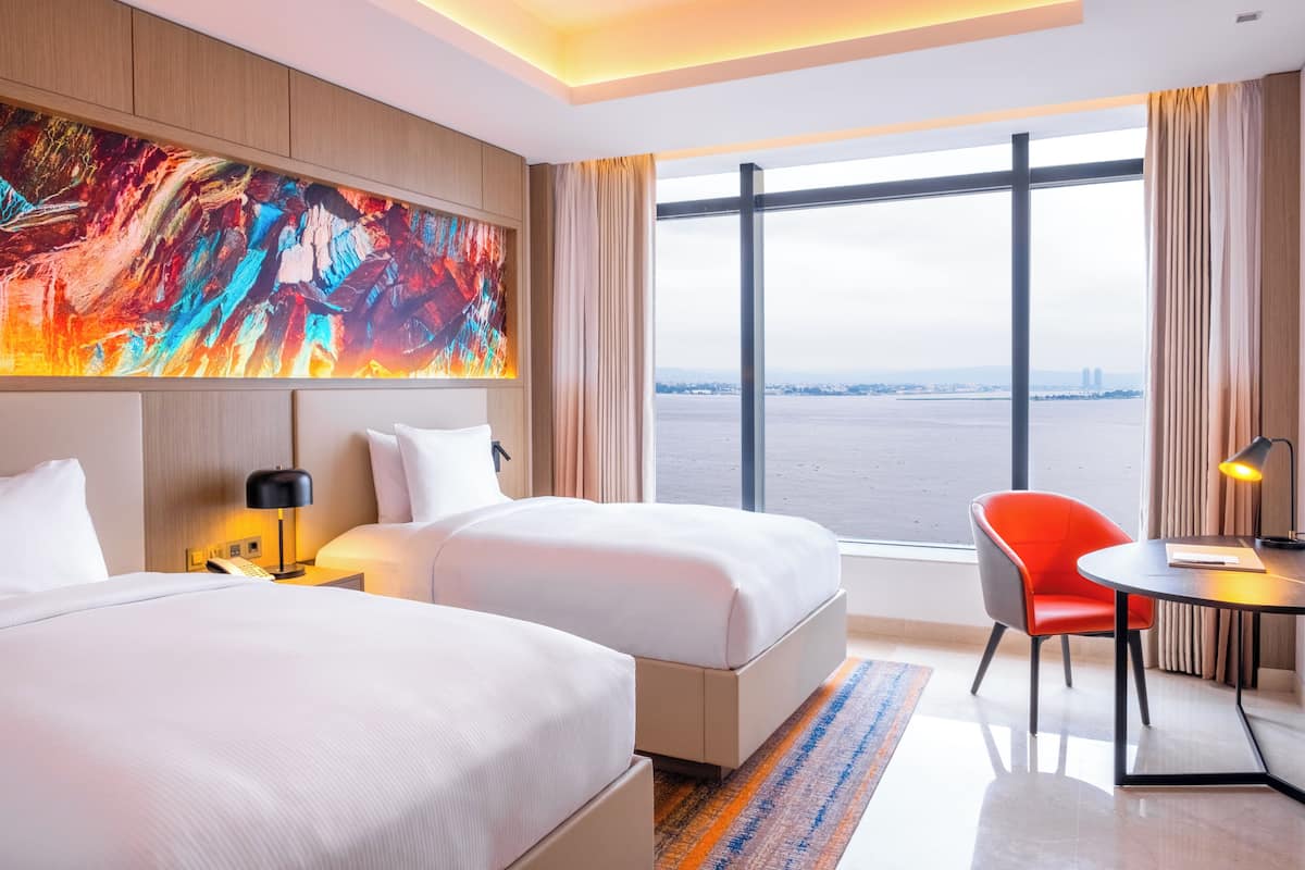 A hotel room with two beds, a large window with a city view, a desk with a chair, and a colorful abstract painting on the wall.