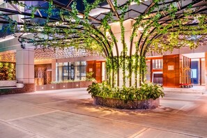 Un moderno vestíbulo de hotel con una jardinera central, paredes de vidrio y un techo con diseño geométrico.