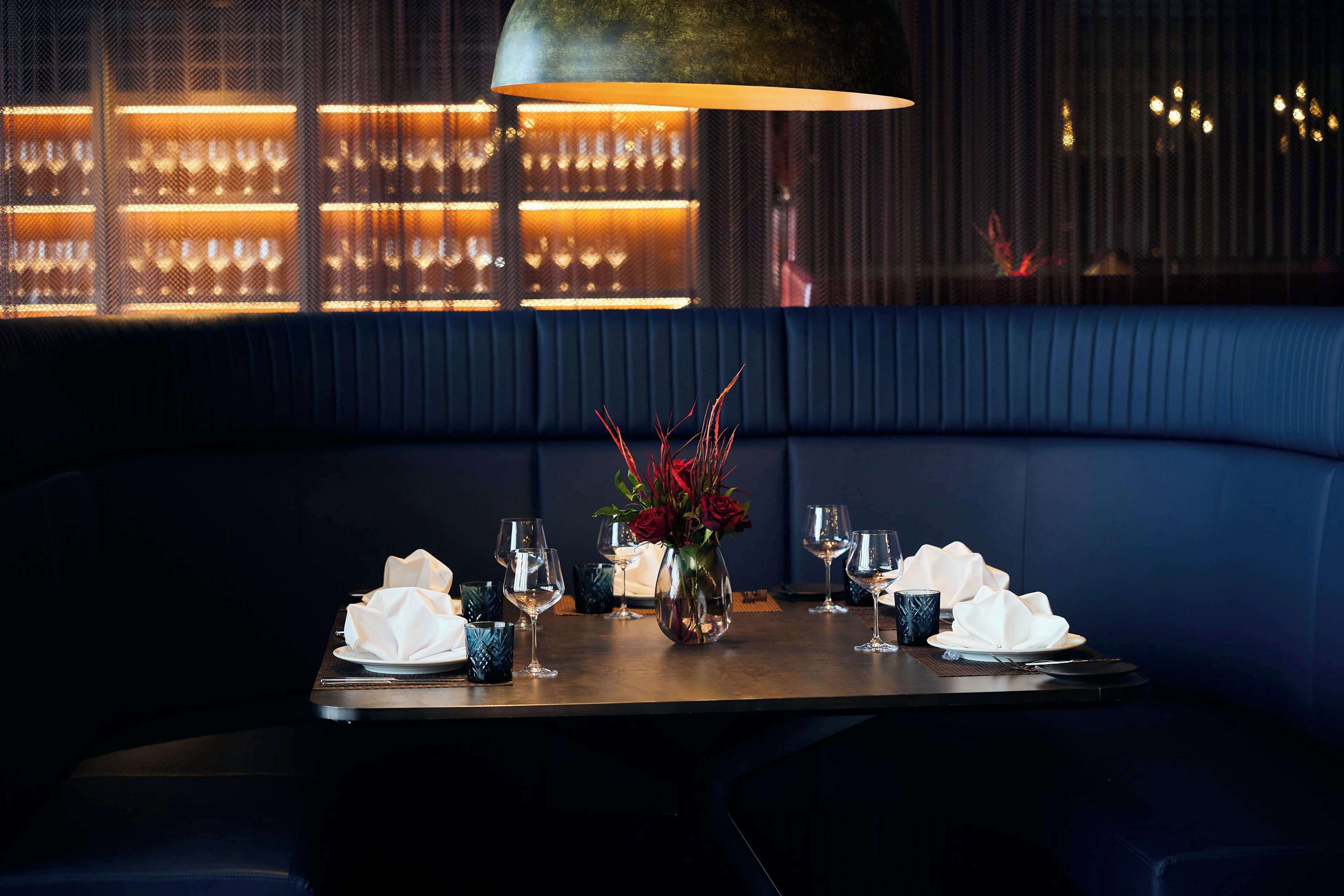 A dark blue booth with a table set for two, featuring a vase with red flowers, wine glasses, and folded napkins.