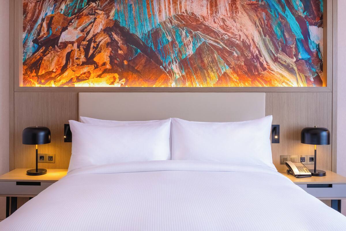 A hotel room with a large bed, two bedside tables with lamps, a phone, and a colorful abstract painting above the bed.