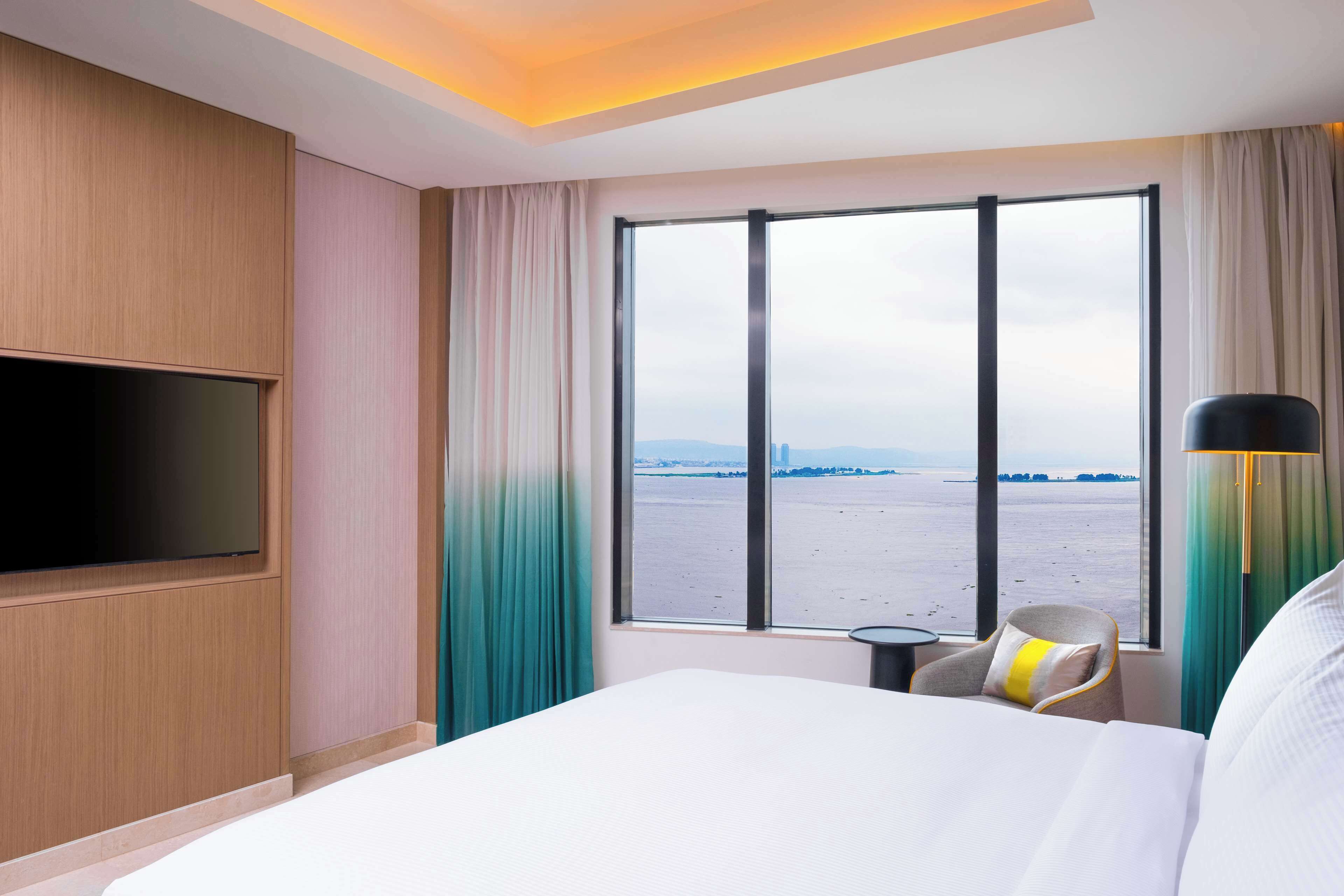 A hotel room with a large bed, a flat-screen TV mounted on the wall, a view of the water, and a chair with a pillow.