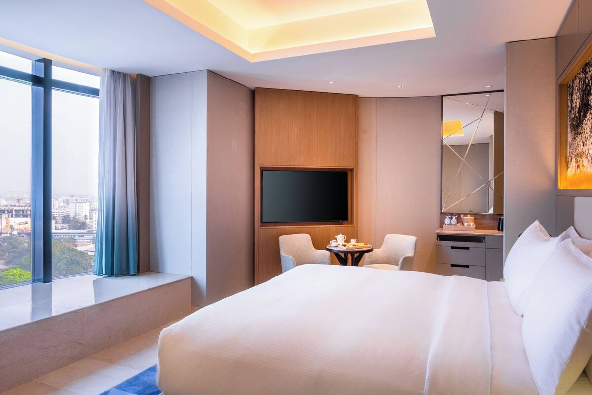 A modern hotel room with a large bed, a TV, a small table with a cup and saucer, and a view of the city through large windows.