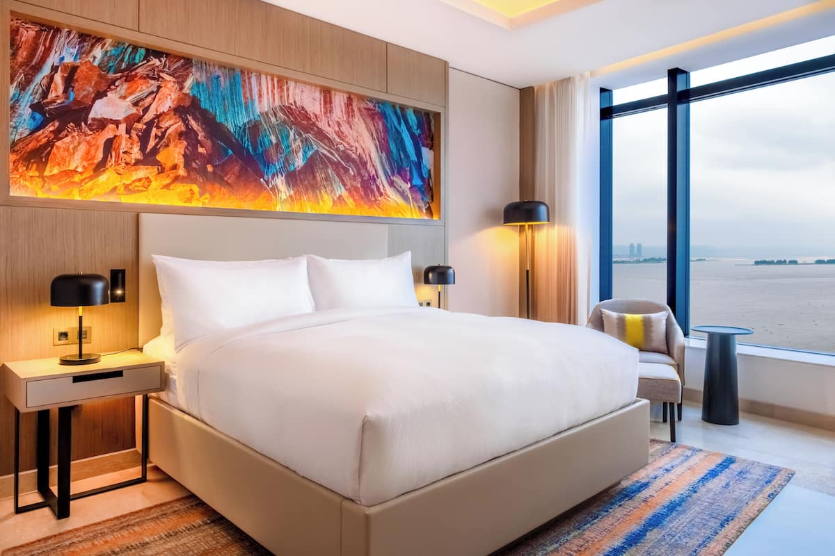 A modern hotel room with a large bed, a nightstand, a desk, a chair, and a large window with a city view.