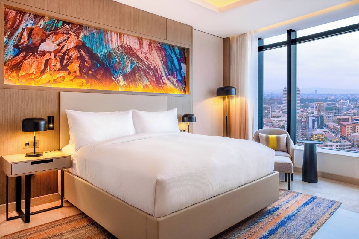 A modern hotel room with a large bed, a nightstand with a lamp, a chair, and a view of the city through large windows.