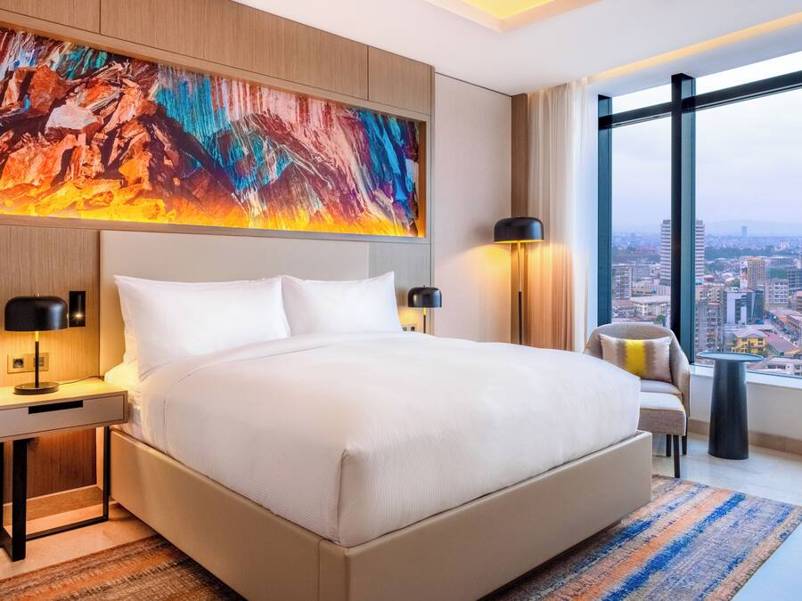 A modern hotel room with a large bed, a nightstand with a lamp, a chair, and a view of the city through large windows.