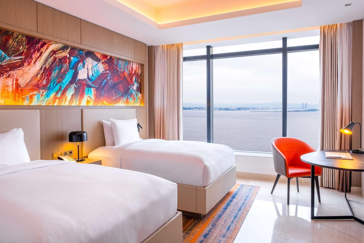 A hotel room with two beds, a large window with a city view, a modern abstract painting, and a small dining area with a chair and table.