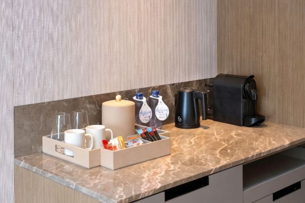 Cocina de habitación de hotel con encimera de mármol, cafetera, hervidor, jarra de café y dispensador de bebidas.