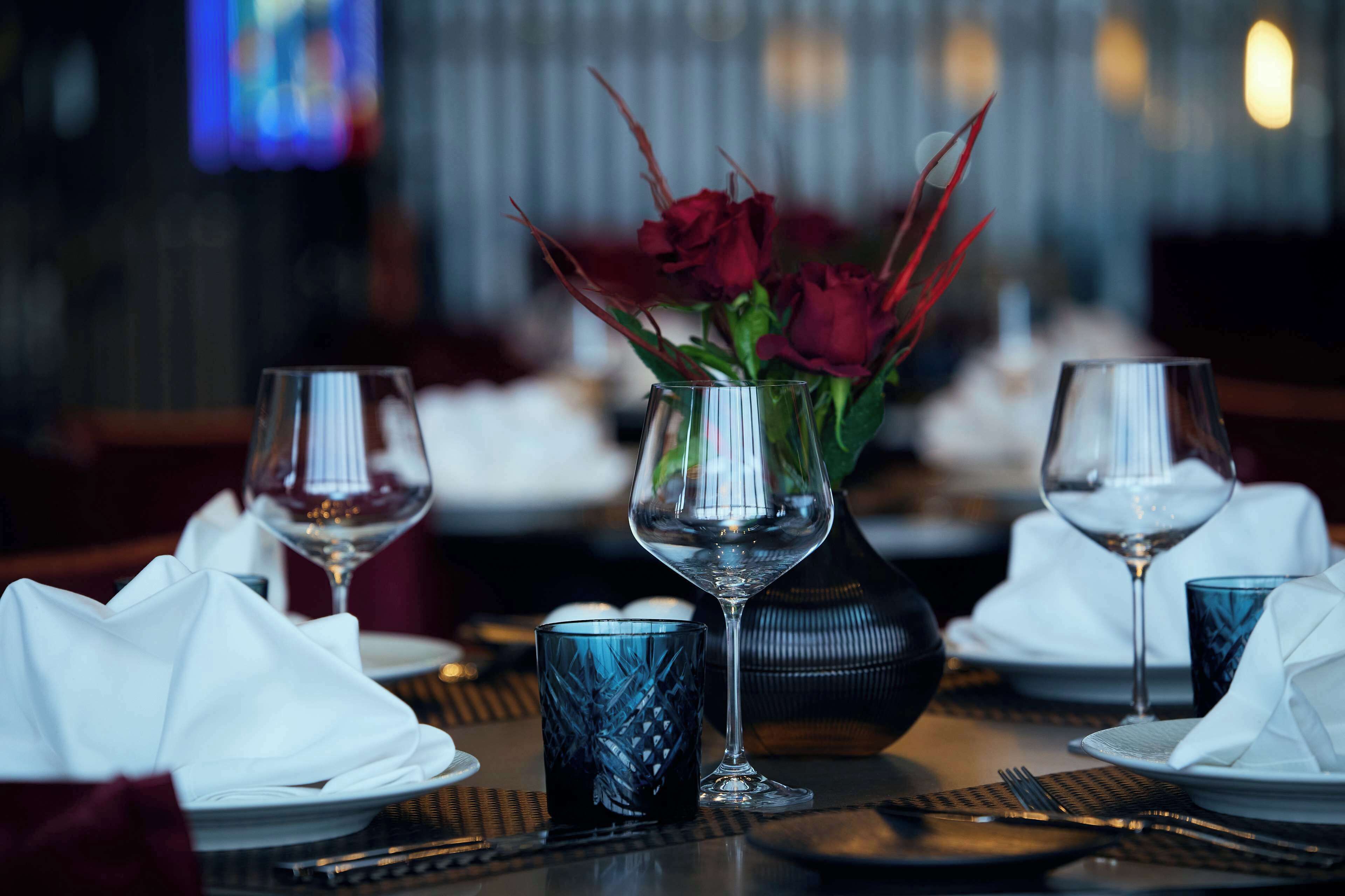 A dining table set with wine glasses, a vase with red roses, and a blue patterned glass.