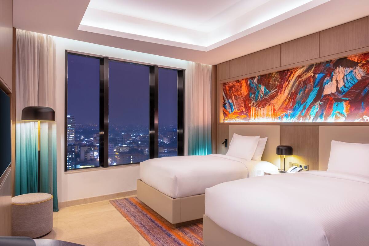 A hotel room with two beds, a large window with curtains, a city view, a colorful abstract painting on the wall, and a small round ottoman.