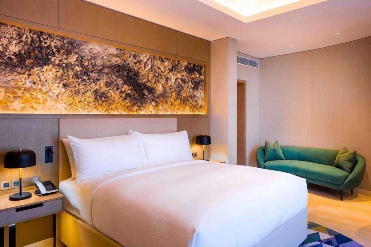 A hotel room with a large bed, bedside lamps, a green sofa, and a large abstract artwork above the bed.